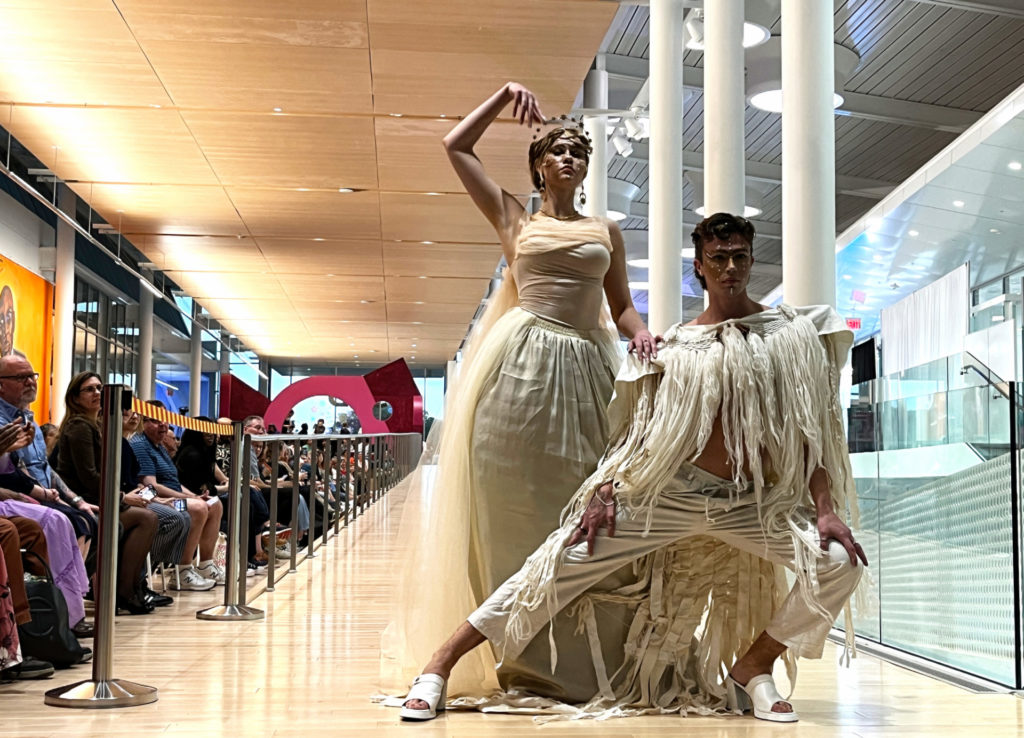Two models pose at ReFashioned 2024. A man and a woman wear off-white and cream colored fabrics. She has a crown on her head and lifts her right arm up in a pose. He wears a top with fringe and tassels, has futuristic makeup on his face, and poses in a lateral lean to his left.