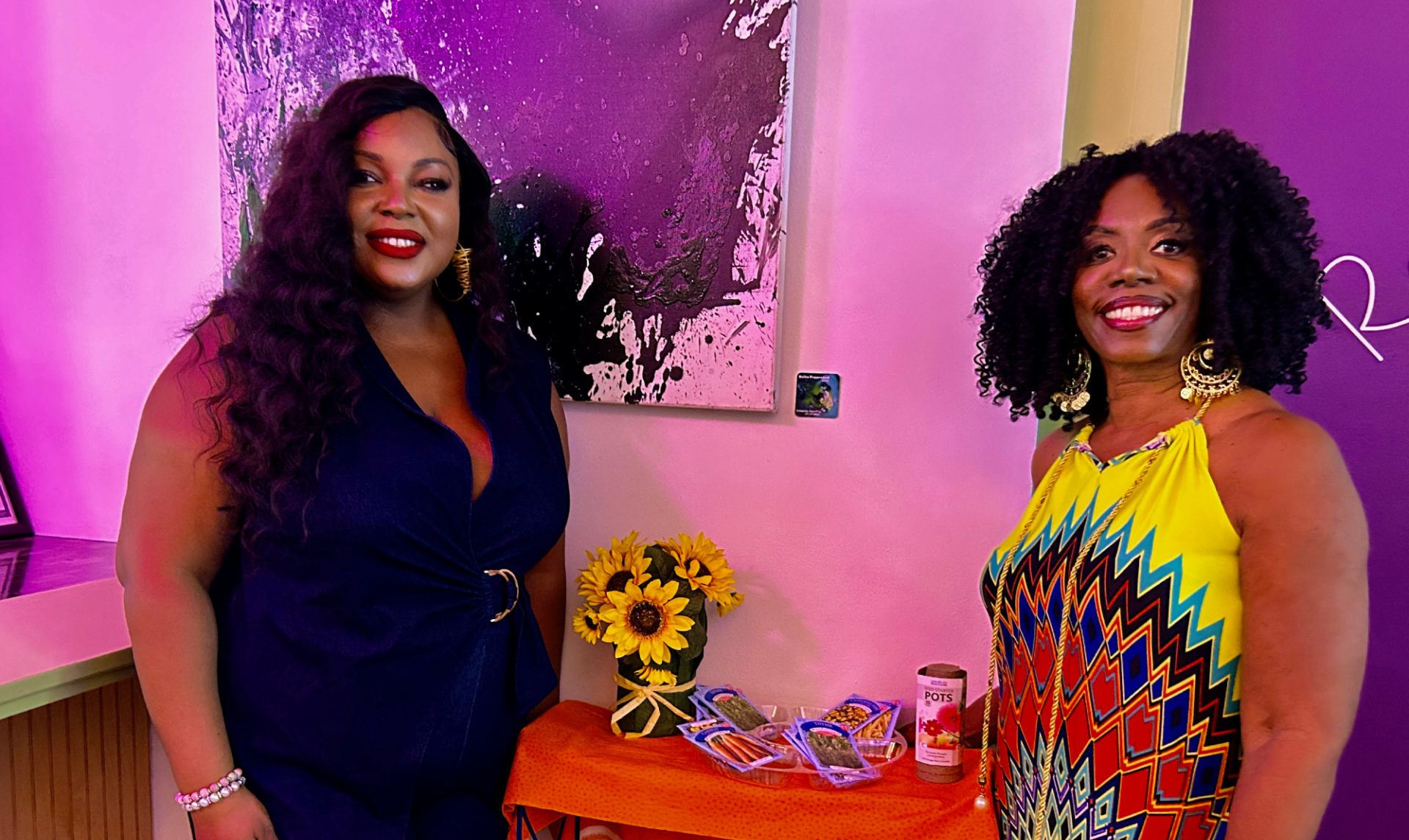 Two Black women stand smiling against a purple and yellow background. Between them is a table with coloring books and crayons and seed packets.