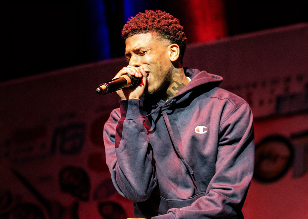 A male performer on stage in a thoughtful pose, holding a microphone. He is dressed in a dark blue hoodie, under soft lighting that creates a contemplative atmosphere.