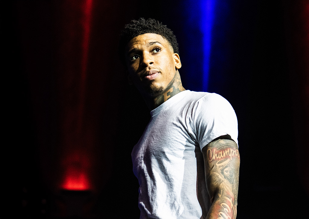 The photograph displays a male performer, up close, as he looks off to the side. He wears a white t-shirt that highlights his tattooed arms. The background is a soft blur of red lighting, focusing attention on his contemplative expression.