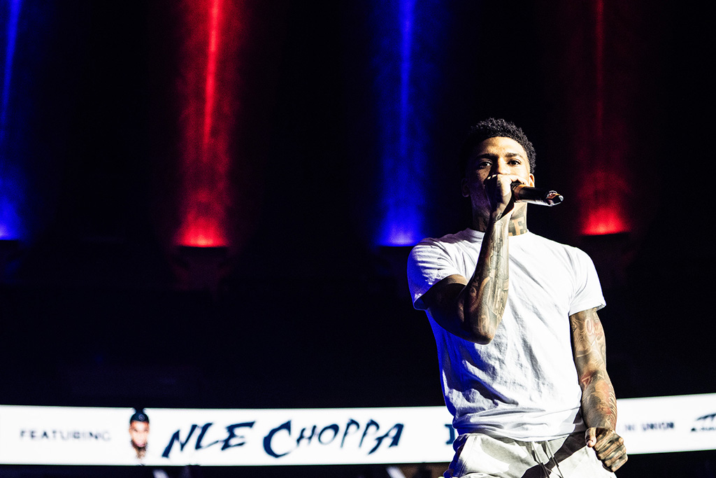 In this vibrant stage scene, a male performer actively engages with the audience, microphone in hand, under dramatic red and blue lighting. His casual white t-shirt contrasts sharply with the dark background, making his animated posture and tattoos stand out.