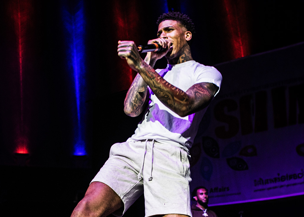 A male performer is captured mid-song, intensely focused, singing into a microphone. He is wearing a white t-shirt and light grey shorts, his arms and neck adorned with tattoos. The stage lighting casts a dramatic red and blue glow, emphasizing his dynamic presence.