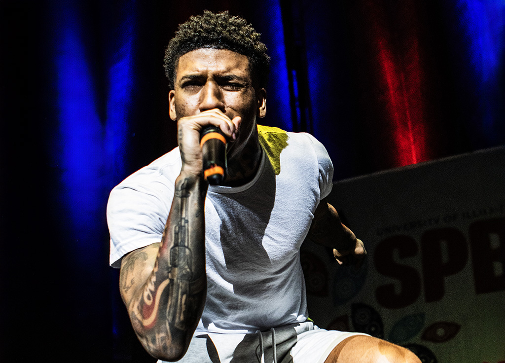 A male performer energetically engaging with the audience, singing into a microphone. He is wearing a white t-shirt layered under a yellow garment, with his tattoos visible on his arms. The stage background glows with intense blue and red lights, enhancing the dynamic expression of his performance.