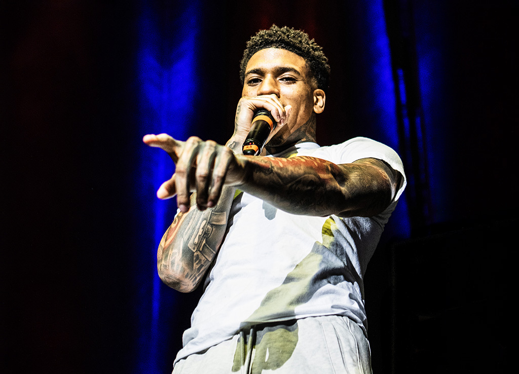 A performer pointing towards the audience, connecting with them as he performs. He is dressed in a white t-shirt, highlighting his tattooed arms. The stage is lit with deep blue and red lights, which adds to the intensity of the scene.