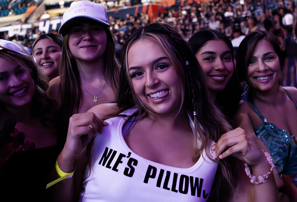 A group of young women at a concert, smiling and enjoying the event. One of them is wearing a white tank top with the text "NLE's Pillow," adding a personal touch to the scene. The background is filled with other concert attendees, creating a lively and joyful atmosphere.