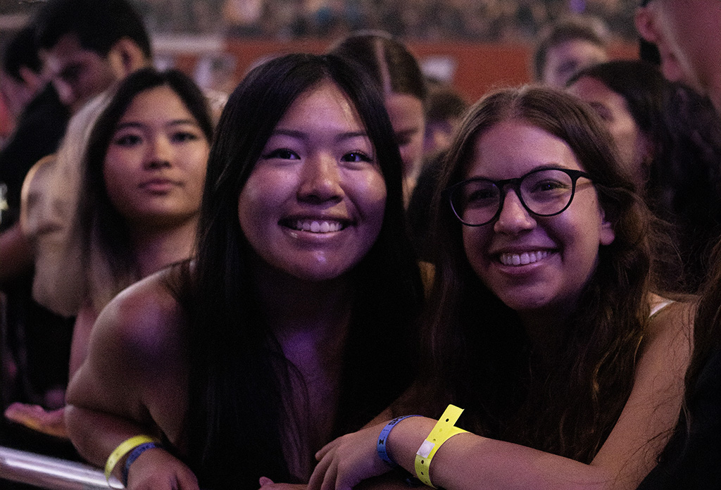Two young women are featured at the forefront of a crowd, smiling broadly as they enjoy a concert. They appear to be in a moment of joy, surrounded by other attendees under the dim lighting of the venue, which adds a warm and inviting feel to the scene.