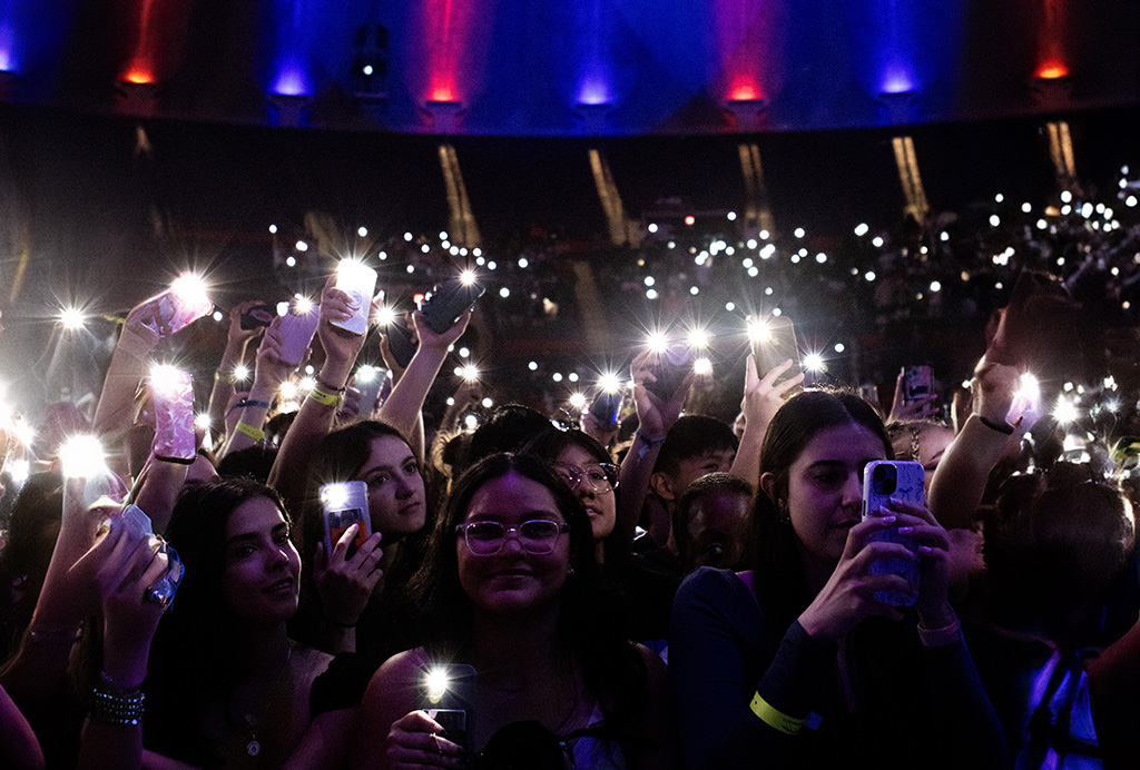 The first image depicts a crowd of concertgoers with their smartphones raised, capturing the moment with lights shining from the screens, creating a sparkling effect throughout the crowd. The atmosphere is vibrant with blue and orange lights illuminating the scene from above, adding a celebratory glow over the audience.