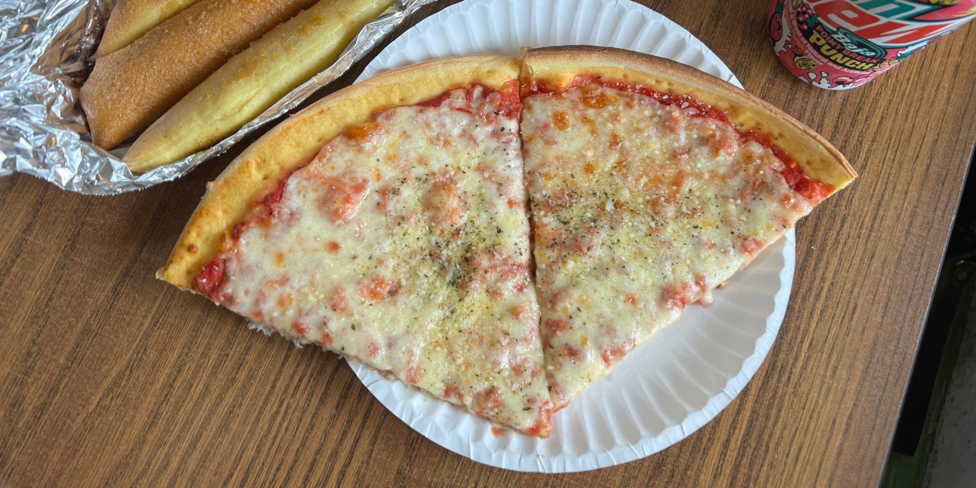 Two slices of thin-crust pizza on a white paper plate beside three breadsticks in tin-foil with a can of Mountain Dew Baja fruit punch.