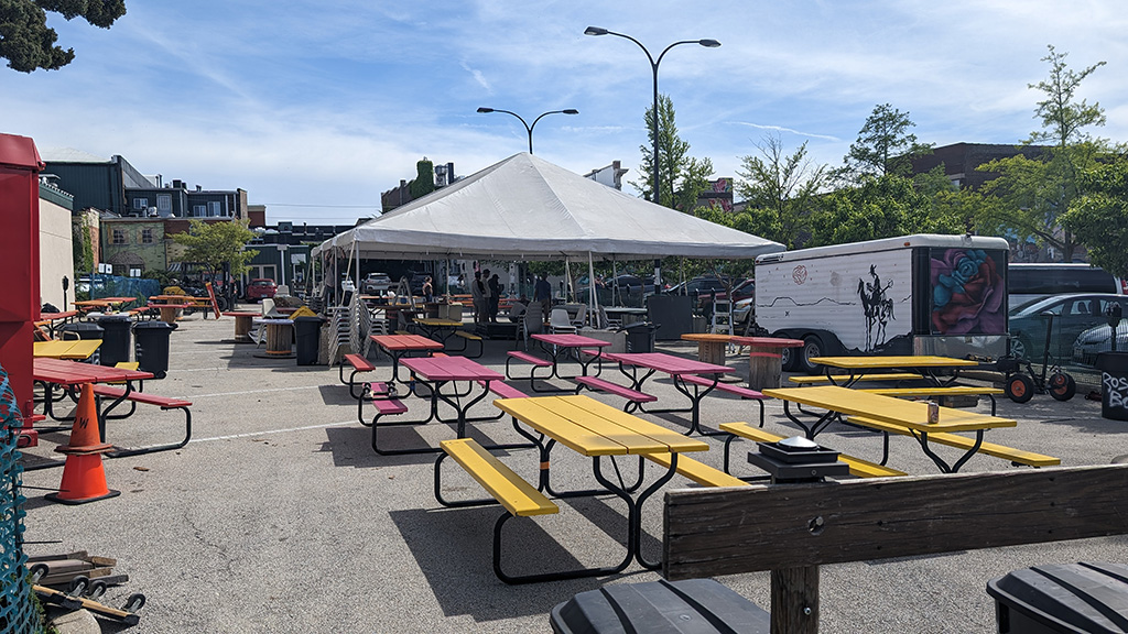 An outdoor setup for an event under a large white tent, surrounded by colorful picnic tables arranged neatly on a parking lot. The scene is set in an urban environment with buildings in the background and several vehicles parked nearby, suggesting a community or public gathering space.