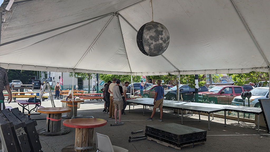 A large outdoor tent setup, possibly for an event, with people gathered underneath. The tent is white, covering a space equipped with several picnic tables and a suspended moon-like decoration, all located in a parking lot.