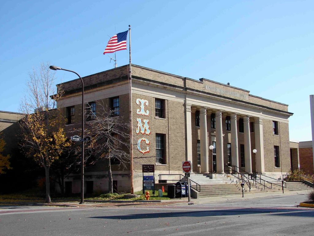 A picture of a large two story gray brick building. It has steps going up and four huge columns. The letters IMC are painted on the front corner and there is an American Flag on the corner. Blue sky is above the building. https://www.facebook.com/photo/?fbid=10152755436614325&set=p.10152755436614325