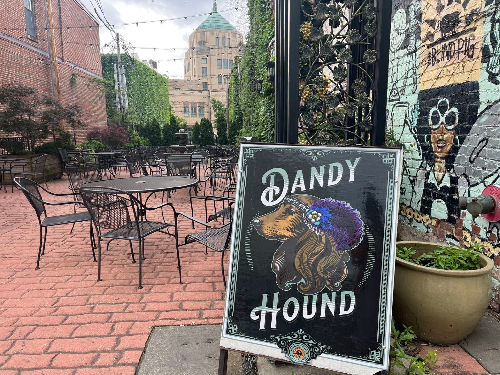 A sign for the Dandy Hound sits against a black archway into a brick paved patio space. In the background is the City of Champaign building and patio lights above the black wire table and chairs.