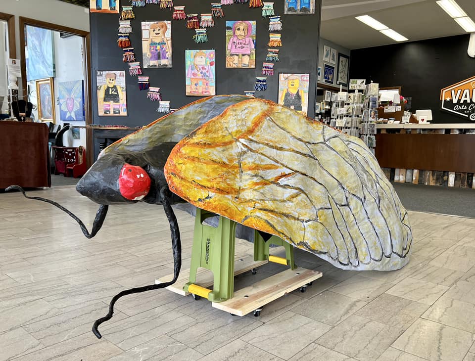 Photo of a paper mache version of a giant cicada. It has a black body and big red eyes with orange and white wings.