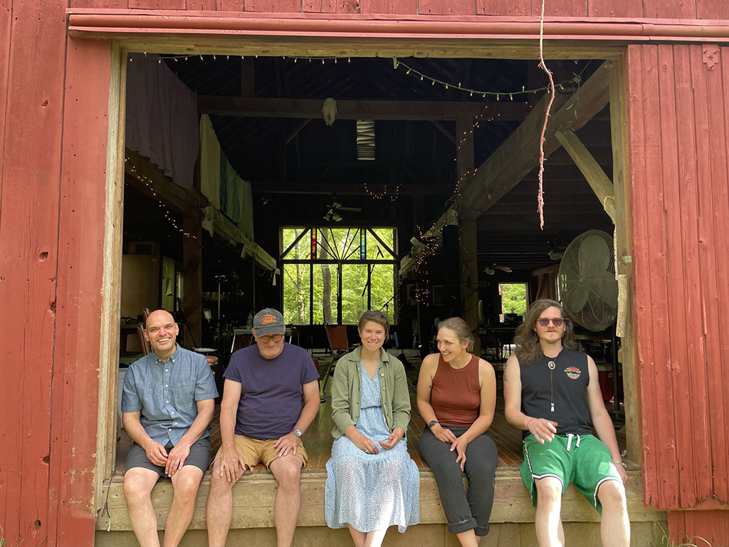 A group of five individuals sits on the edge of a red barn's loading dock, immersed in a casual group interaction. They are dressed for comfort and warmth, suggesting a friendly gathering or a break during a collaborative event. The barn's large open doors and windows show a glimpse of the musical equipment inside, indicating a creative or musical setting.