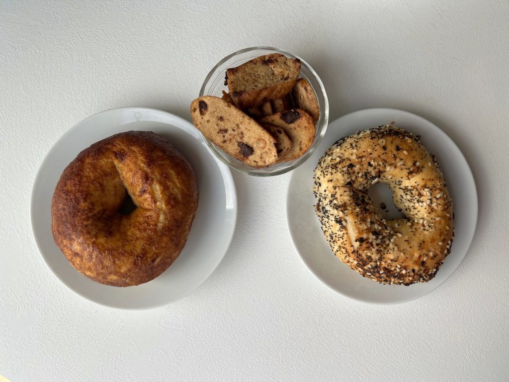 Two white plates have bagels (one cinnamon sugar, one everything) and a glass bowl of bagel chips with chocolate chips.