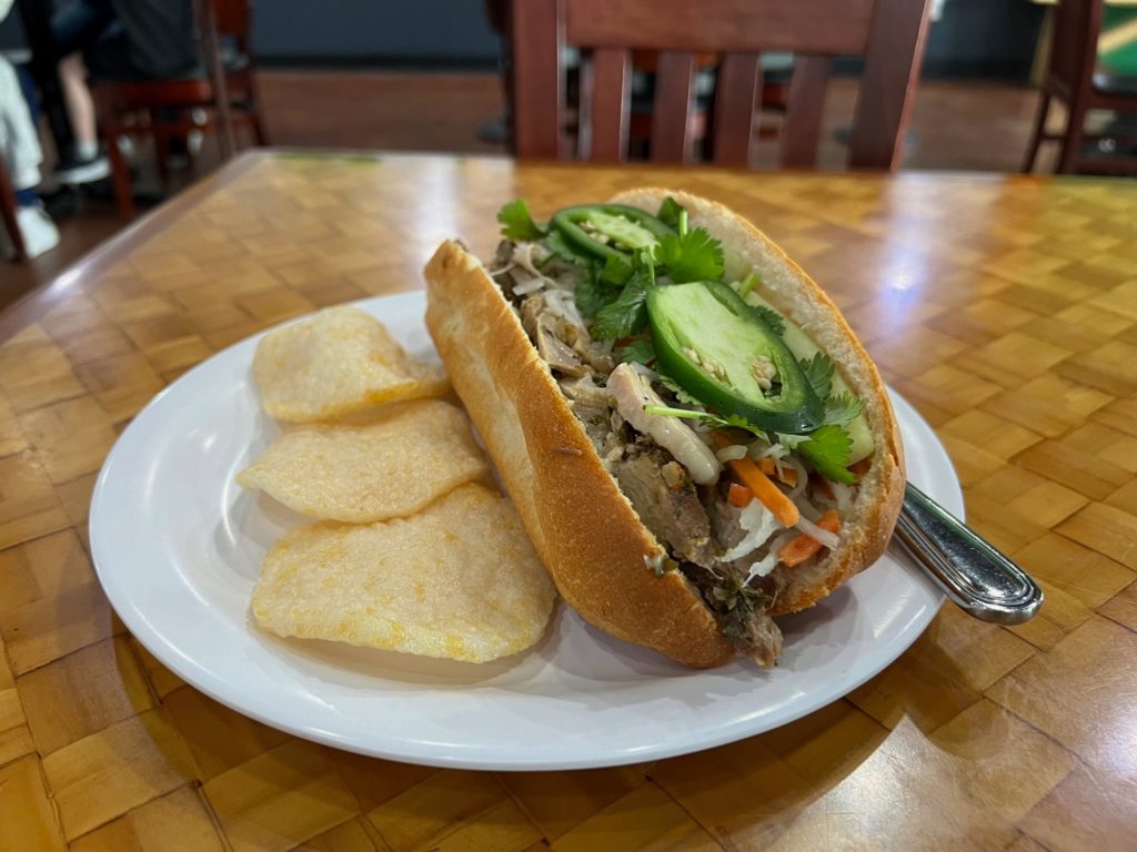 A banh mi baguette with Italian pork and banh mi toppings with three shrimp chips and a metal knife on a white plate.