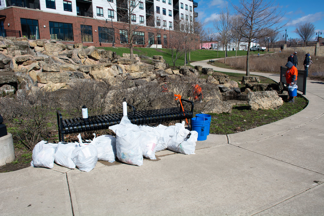 View of a park bench against which are at least nine filled white trash bags. There are buckets and trash pickers nearby, and a volunteer trash collector in an orange t-shirt on the right of the image.