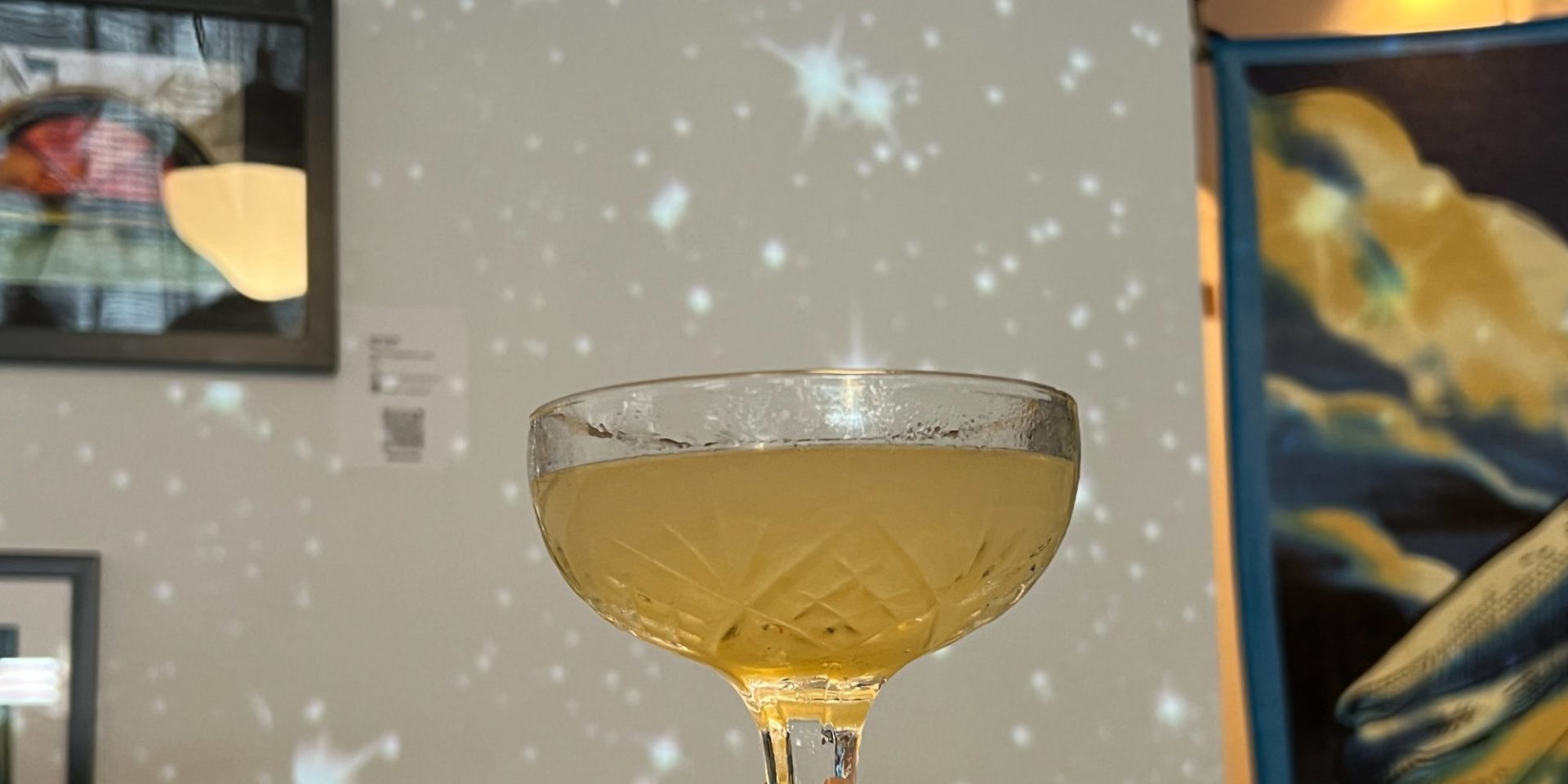A coupe glass with a golden yellow drink in front of a white wall with projected starbursts.