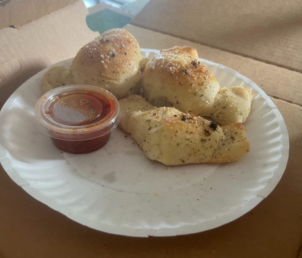Three garlic knots with a side of marinara in closed takeout cup on a paper plate.
