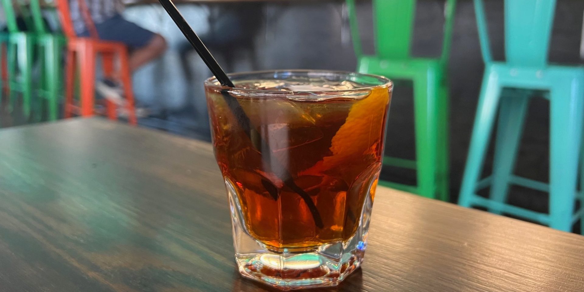 A rocks glass with an old-fashioned and an orange peel with a black straw on a wooden table inside The Space restaurant in Downtown Champaign.