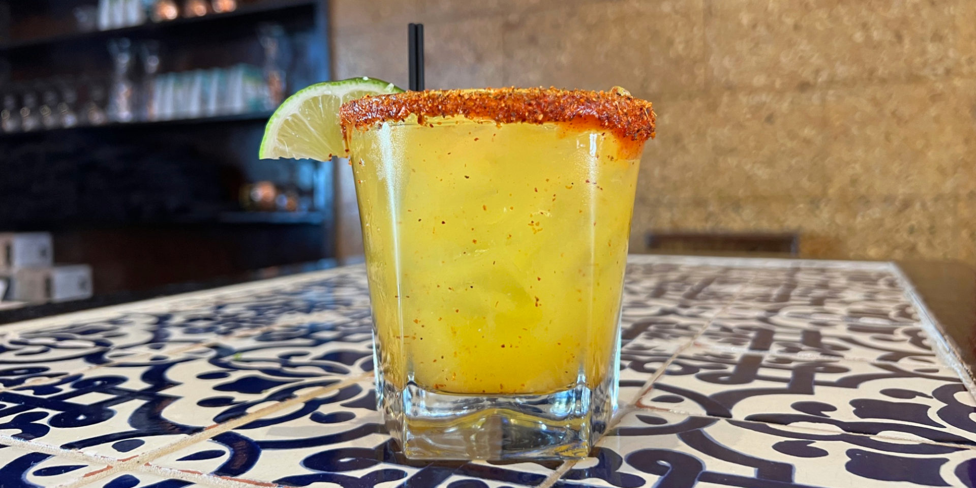 A yellow margarita with a red tajin rim and a slice of lime on a clear rocks glass with two skinny black straws.