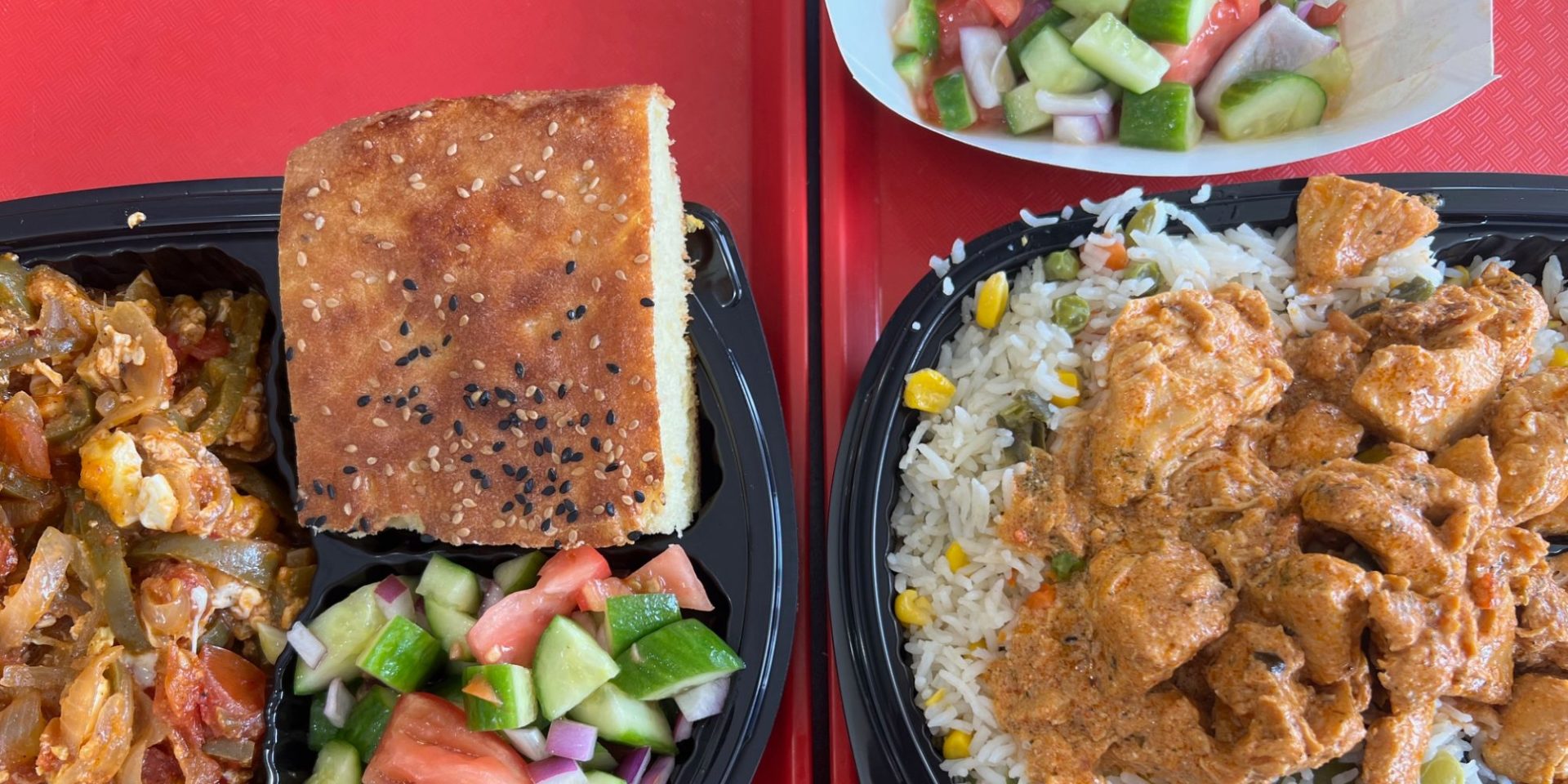 On two red trays, two black plastic compartment-takeout containers have North African Cuisine: shakshouka with salad and bread, the other rice with vegetables topped with sauced chicken.