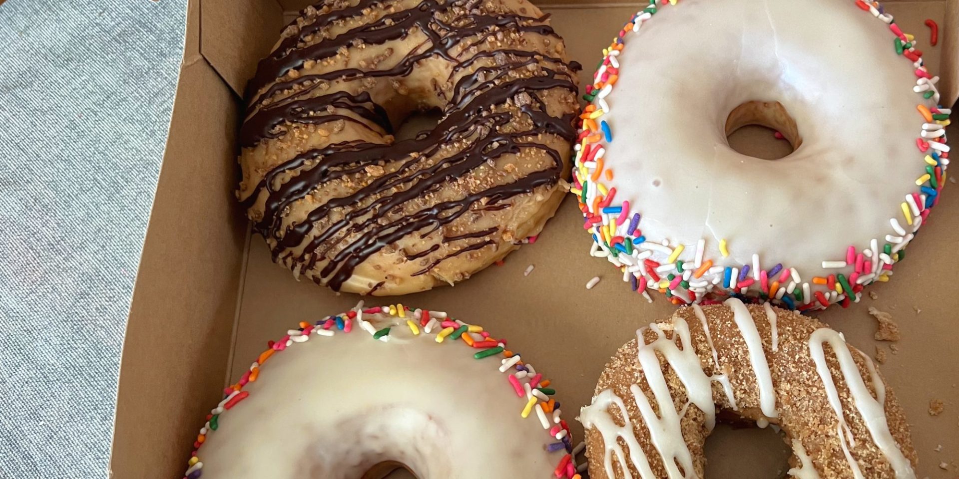 Four doughnuts in a brown box: one chocolate drizzled, two vanilla with rainbow sprinkles along the circumference, and a white chocolate drizzled coffee cake doughnut.
