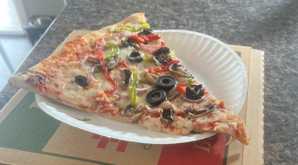 Veggie supreme slice on a white paper plate on a brown pizza box.