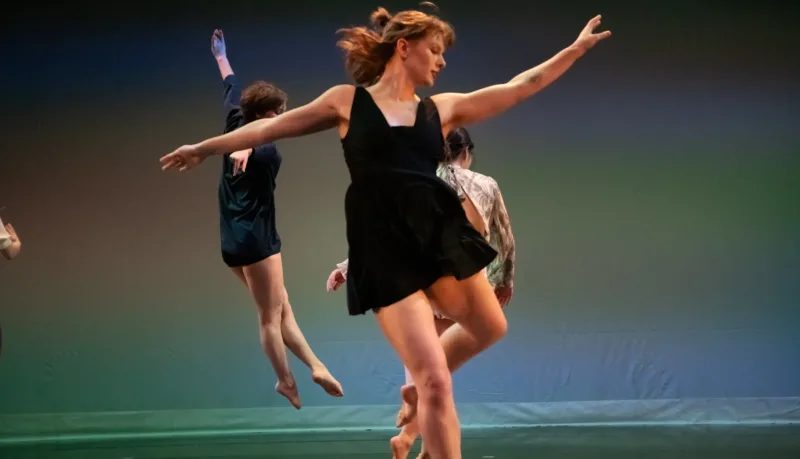 A woman in a black dress dancing, two people dancing behind her facing away against a blue and green backdrop