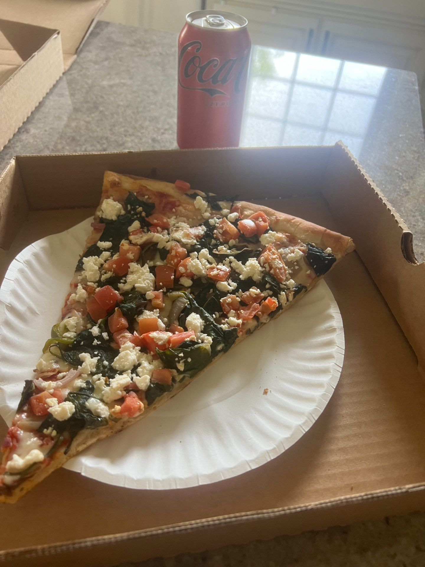 A single slice of spinach pizza on a white paper plate inside a pizza box with an unopened can of Coca Cola zero.