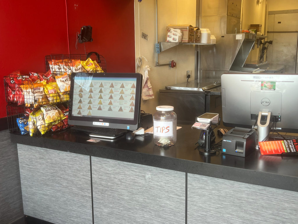 The interior of the Savoy location for Vinny's Pizza By The Slice has a counter with individual chips, a kiosk touch screen, a clear jar for tips, and a cashier stand.