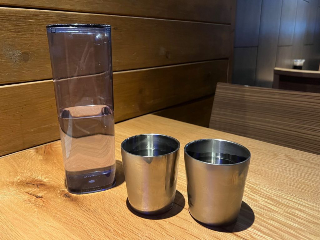 A clear pitcher of water beside two metal cups of water.