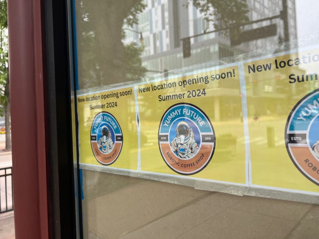 A window has repeated yellow signs reading "New location opening soon! Summer 2024" for robot coffee shop Yummy Future.